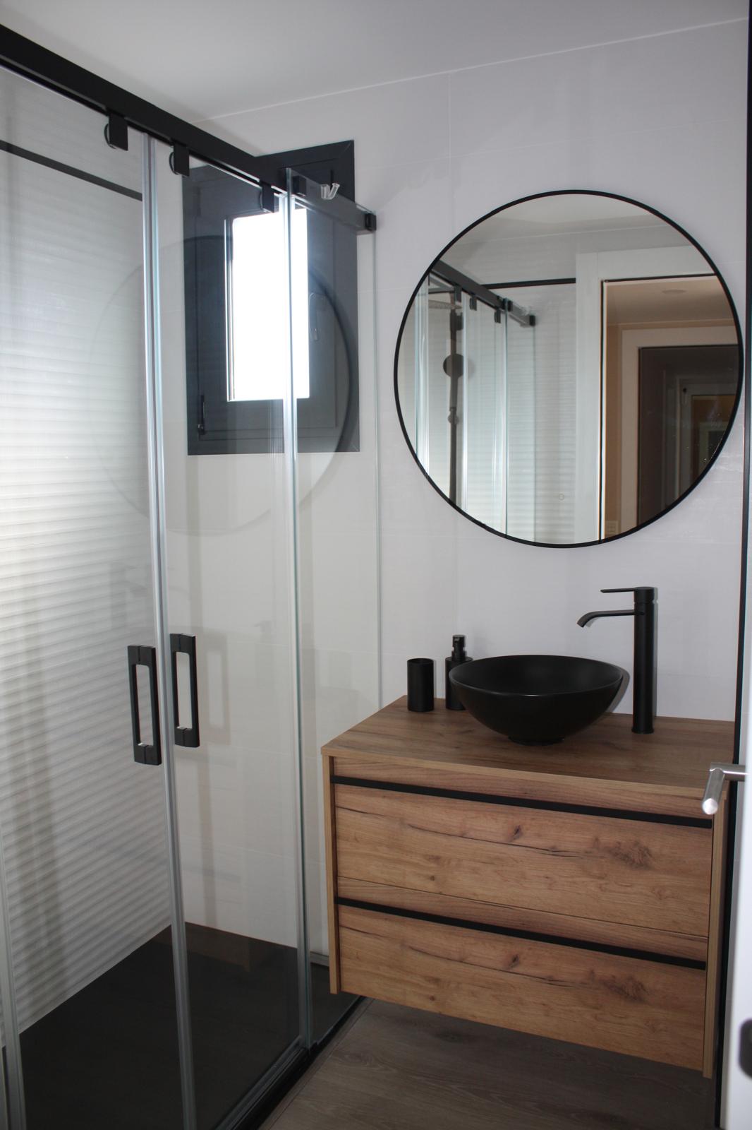 Baño con grifería negra mate, lavabo sobre encimera de madera y espejo circular